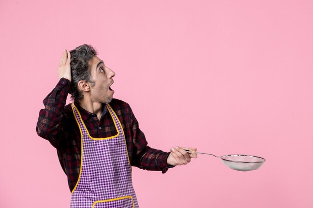 vista frontal jovem macho na capa segurando peneira no fundo rosa trabalhador cozinheiro profissão comida cor cozinha marido horizontal