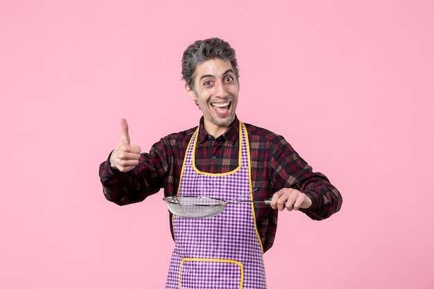 vista frontal jovem masculino na capa com peneira e colher no fundo rosa cozinheiro horizontal comida profissão marido cozinha uniforme trabalhador