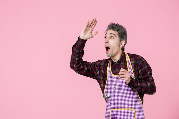 vista frontal jovem masculino na capa segurando uma colher de sopa apontando para algo rosa fundo cozinheiro cozinha cozinha comida profissão marido horizontal