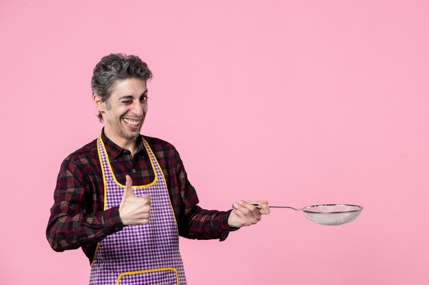 vista frontal jovem masculino na capa segurando uma peneira no fundo rosa trabalhador cozinheiro trabalho profissão comida cozinha marido horizontal