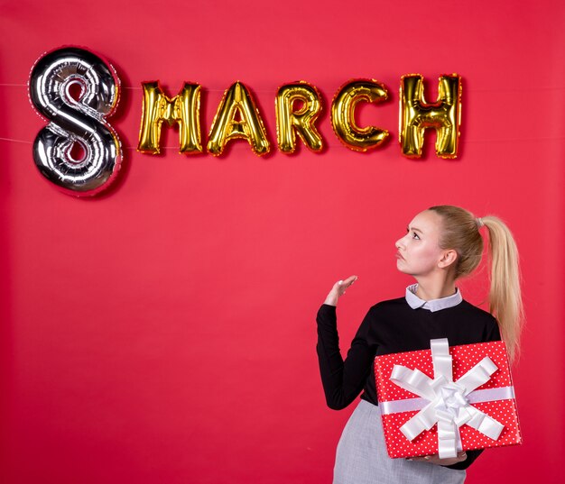 Vista frontal jovem mulher segurando presente com decoração de marcha em fundo vermelho paixão salão mulher dia igualdade feminino dinheiro compras