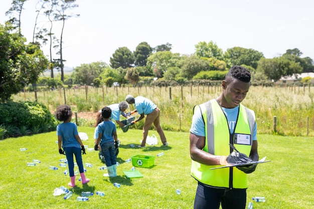 Vista frontal de un joven afroamericano de pie en un campo con un chaleco y guantes de alta visibilidad, escribiendo en un portapapeles, mientras que un grupo diverso de voluntarios recoge basura y recicla en el backgr