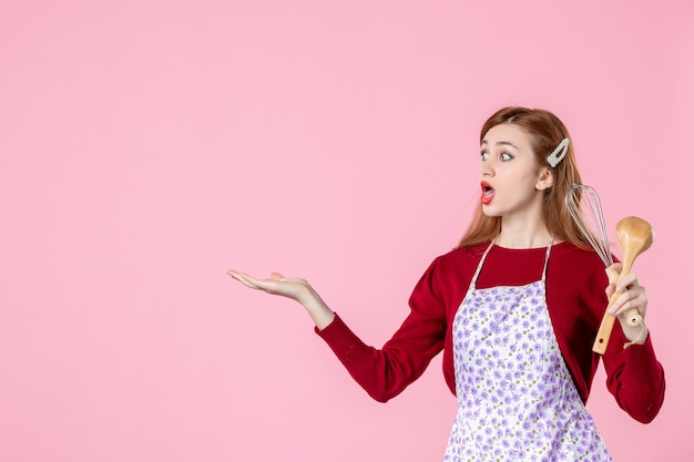 Vista frontal joven ama de casa posando con un batidor y una cuchara de madera sobre fondo de color rosa pastel de pastel de mujer profesión masa uniforme cocina horizontal