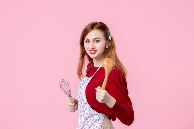 Vista frontal joven ama de casa posando con un batidor y una cuchara de madera sobre fondo rosa cocinando masa pastel horizontal profesión mujer pastel cocina