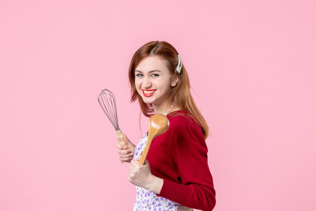 Vista frontal joven ama de casa posando con un batidor y una cuchara de madera sobre fondo rosa cocinando masa pastel horizontal profesión mujer pastel uniforme de cocina