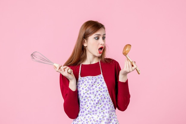 Vista frontal joven ama de casa posando con batidor y cuchara de madera sobre fondo rosa pastel profesión cocinando masa tartas horizontales uniforme mujer cocina