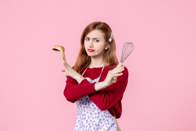 Vista frontal joven ama de casa posando con un batidor y una cuchara de madera sobre fondo rosa profesión cocinando masa mujer pastel cocina uniforme pastel horizontal