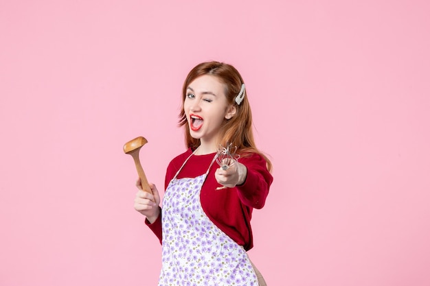 Vista frontal joven ama de casa sosteniendo un batidor y una cuchara de madera sobre fondo rosa cocina cocina mujer pastel masa de tarta uniforme horizontal