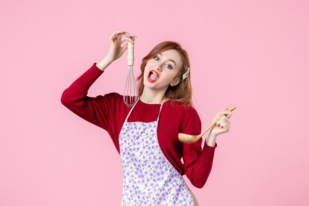 Vista frontal joven ama de casa sosteniendo un batidor y una cuchara de madera sobre fondo rosa cocina cocina uniforme profesión mujer pastel horizontal