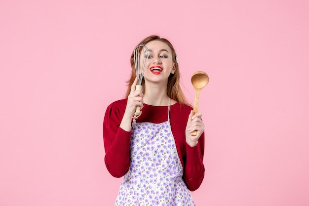 Vista frontal joven ama de casa sosteniendo un batidor y una cuchara de madera sobre fondo rosa comida cocina pastel mujer compras cocina cocina pastel