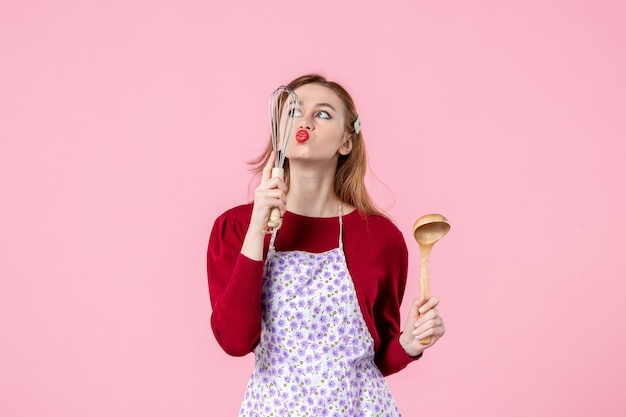 Vista frontal joven ama de casa sosteniendo un batidor y una cuchara de madera sobre fondo rosa comida cocina pastel mujer compras dinero cocina cocinando pastel