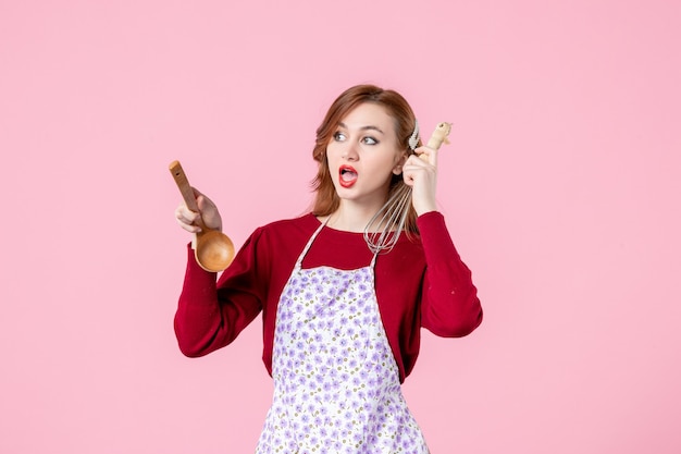 Vista frontal joven ama de casa sosteniendo un batidor y una cuchara de madera sobre fondo rosa mujer cocinando pastel de tarta horizontal profesión cocina