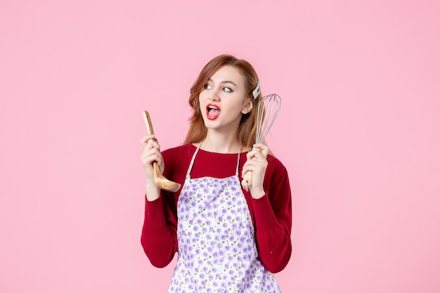 Vista frontal joven ama de casa sosteniendo un batidor y una cuchara de madera sobre fondo rosa pastel horizontal mujer cocina uniforme profesión cocina masa para tartas