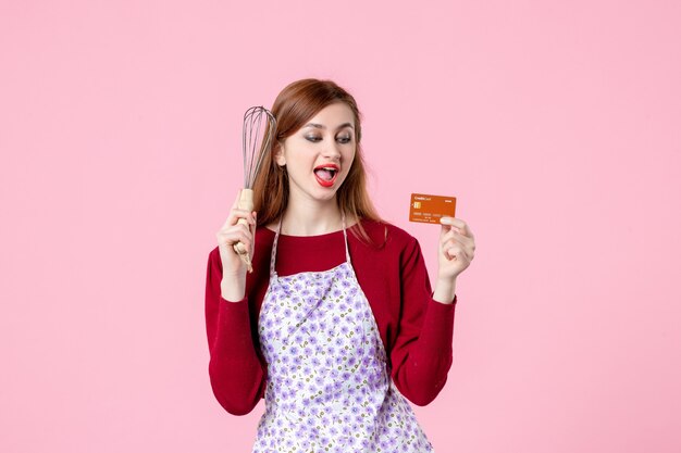 Vista frontal joven ama de casa sosteniendo un batidor y una tarjeta bancaria sobre fondo rosa pastel de cocina pastel mujer compras dinero cocina alimentos colores cocinar