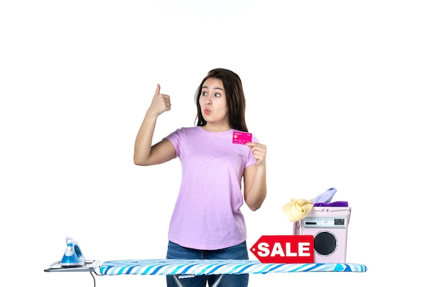 Foto vista frontal joven ama de casa con tarjeta bancaria rosa sobre fondo blanco color de ropa de trabajo de lavandería compras quehaceres domésticos venta dinero de hierro