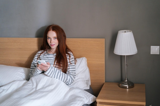 Vista frontal de una joven atractiva que usa teléfono móvil, escribe un mensaje en línea en las redes sociales, mira la cámara, se sienta en una cama acogedora por la noche en el dormitorio.