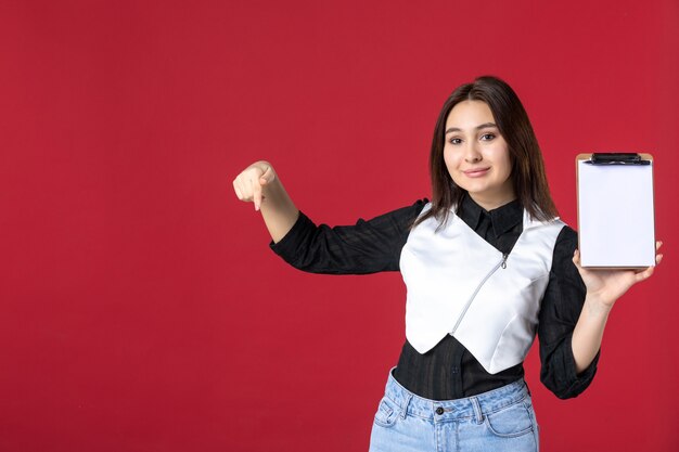 Vista frontal joven bonita camarera en uniforme sosteniendo nota de archivo para pedidos sobre fondo rojo trabajo belleza mujer trabajo trabajador noche de comida