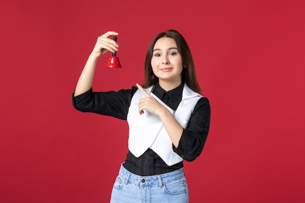 Vista frontal joven camarera en uniforme sosteniendo campanita sobre fondo rojo cena trabajo por la noche trabajo café restaurante mujer belleza color trabajador