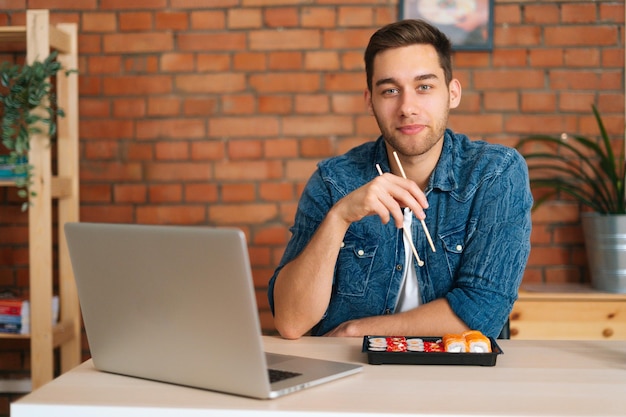 Vista frontal de un joven caucásico feliz comiendo rollos de sushi con palillos sentados en el escritorio con una laptop en la oficina mirando la cámara
