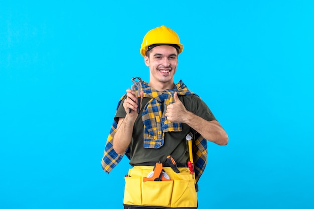 Vista frontal joven constructor masculino con diferentes herramientas sobre fondo azul trabajo plano trabajador constructor edificio arquitectura casa