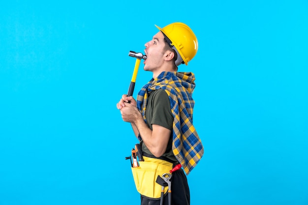 Vista frontal joven constructor masculino sosteniendo un martillo sobre el fondo azul trabajador edificio arquitectura constructor trabajo plano