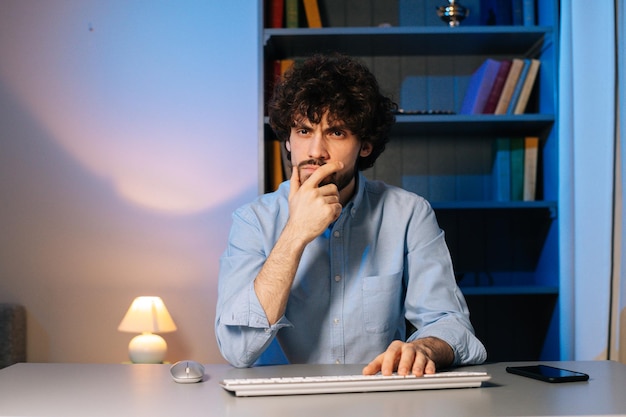 Vista frontal de un joven descontento escribiendo un mensaje en línea en un teclado inalámbrico. Punto de vista de acariciar la barba cuidadosamente mientras se trabaja en la computadora en casa. Concepto de trabajo remoto.