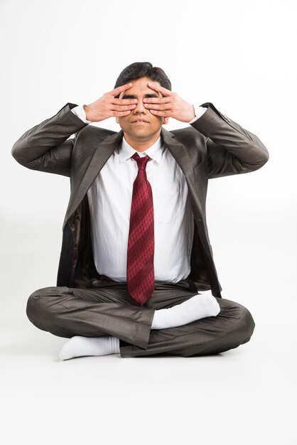 Vista frontal de un joven empresario asiático indio meditando o practicando yoga o pranayama o ejercicio de respiración en traje corporativo en la oficina o aislado sobre fondo blanco.