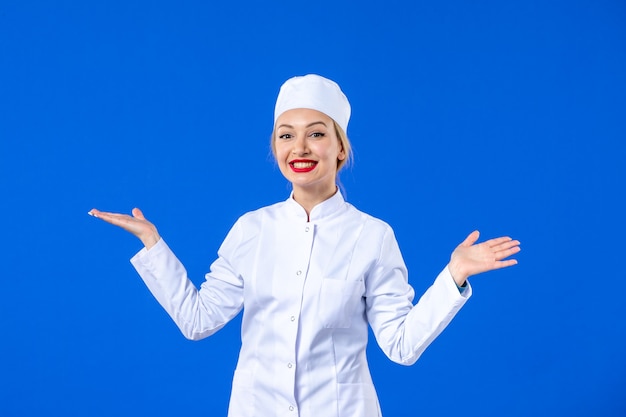 Vista frontal de la joven enfermera coqueta en traje médico blanco sobre fondo azul enfermedad pandémica covid- medicina enfermedad de la salud hospitalaria de medicamentos