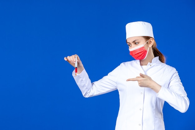 Vista frontal joven enfermera en traje médico blanco con máscara roja e inyección en sus manos en la pared azul