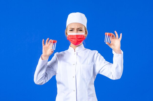 Vista frontal joven enfermera en traje médico con máscara roja sosteniendo frascos sobre fondo azul médico de salud de drogas covid- medicina hospital virus