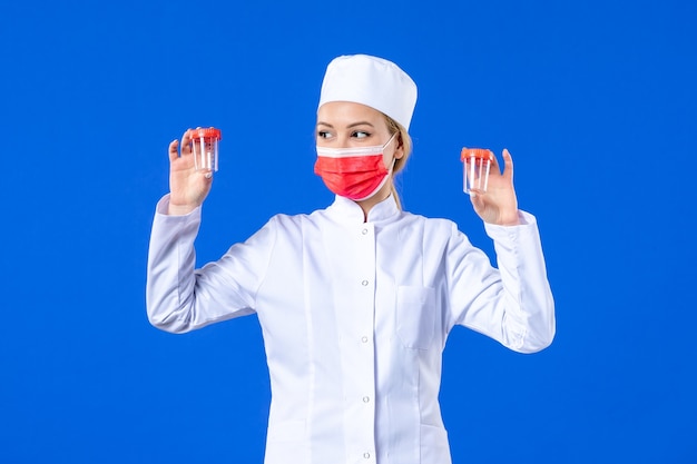 Vista frontal joven enfermera en traje médico con máscara roja sosteniendo frascos sobre fondo azul pandemia de virus de salud covid- doctor de drogas del hospital