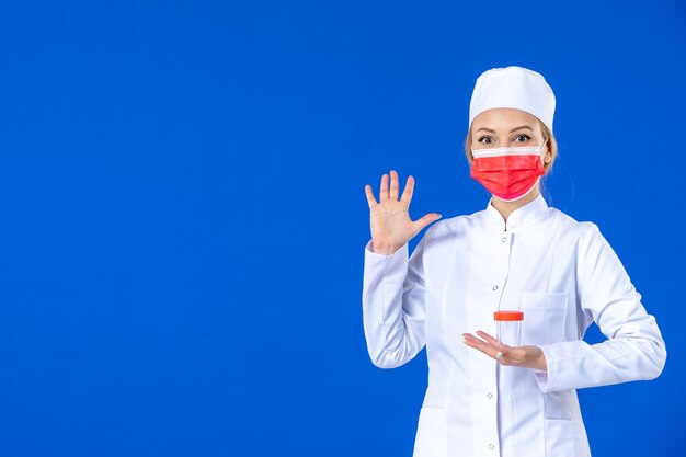 Vista frontal joven enfermera en traje médico con máscara sosteniendo matraz sobre fondo azul hospital virus covid- vacuna pandemia de medicamentos de salud