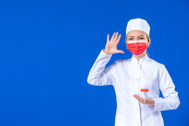 Vista frontal joven enfermera en traje médico con máscara sosteniendo matraz sobre fondo azul médico de salud de drogas hospital covid- medicina de vacuna pandémica