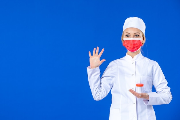 Vista frontal joven enfermera en traje médico con máscara sosteniendo matraz sobre fondo azul médico de salud de drogas hospital covid- medicina de virus de vacuna pandémica