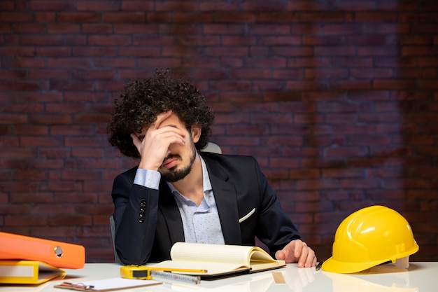 Vista frontal del joven ingeniero sentado detrás del lugar de trabajo en traje contratista de negocios en el interior constructor de trabajo corporativo