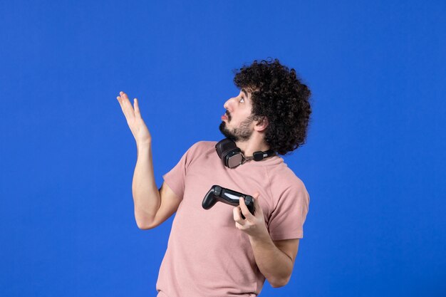 Vista frontal joven jugando videojuegos con gamepad negro sobre fondo azul adolescente virtual sofá juvenil alegría adulta jugador de fútbol ganar