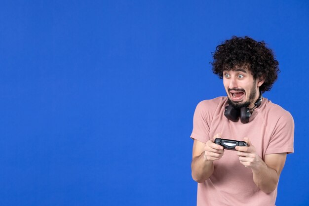 Vista frontal joven jugando videojuegos con gamepad negro sobre fondo azul alegría juvenil adolescente jugador ganador de fútbol sofá virtual