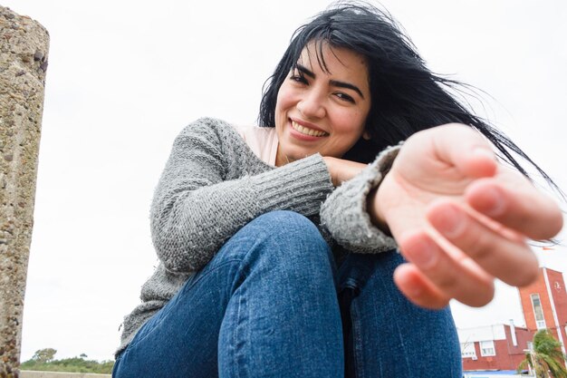 Vista frontal de una joven latina al aire libre sentada feliz con el brazo extendido mirando a la cámara