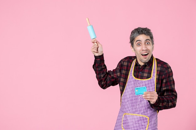 Vista frontal joven macho en cabo sosteniendo pequeño rodillo y tarjeta bancaria sobre fondo rosa trabajo cocina marido trabajador uniforme profesión cocina dinero horizontal