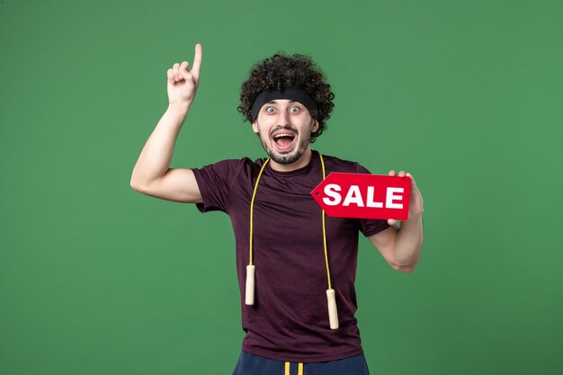 Vista frontal joven macho sosteniendo rojo placa de venta sobre fondo verde atleta gimnasio compras deporte régimen de entrenamiento color de salud