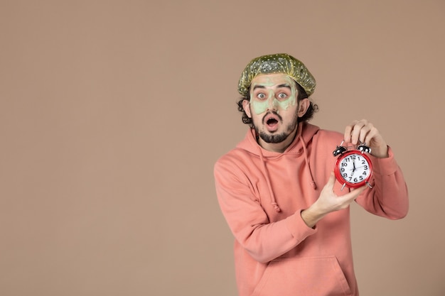 Vista frontal joven con máscara sosteniendo relojes sobre fondo marrón tiempo de spa salón de piel cuidado corporal facial