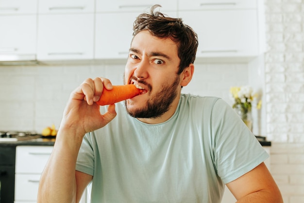 Vista frontal de un joven mordiendo una zanahoria cruda con una expresión feroz e infeliz comiendo sano