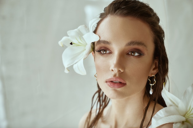 Vista frontal de la joven mujer morena con maquillaje perfecto y rostro fuerte sosteniendo flor de lirio detrás de la oreja