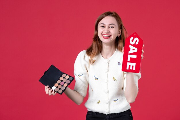 vista frontal joven mujer sosteniendo caja de polvos y rojo placa de venta sobre fondo rojo belleza colores sensual mujer marzo maquillaje piel moda labios compras