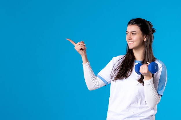 Vista frontal joven mujer sosteniendo mancuernas azul sobre pared azul