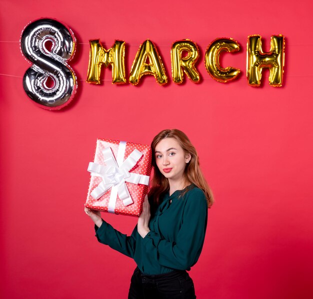 Vista frontal joven mujer sosteniendo presente con decoración de marzo sobre fondo rojo moda amor mujer día de la mujer regalo femenino feliz glamour vacaciones