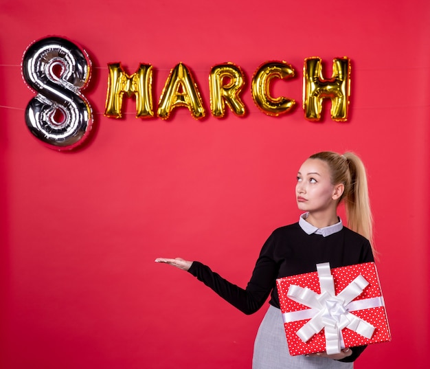 Vista frontal joven mujer sosteniendo presente con decoración de marzo sobre fondo rojo salón de pasión día de la mujer lujoso igualdad compras dinero