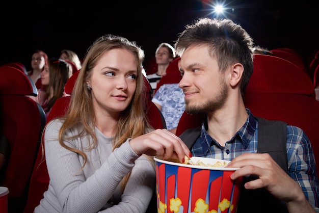 Vista frontal de la joven pareja comiendo palomitas de maíz y mirándose cara a cara durante la comedia en el cine. Chica rubia y guapo con cita romántica y disfrutar de una película divertida.