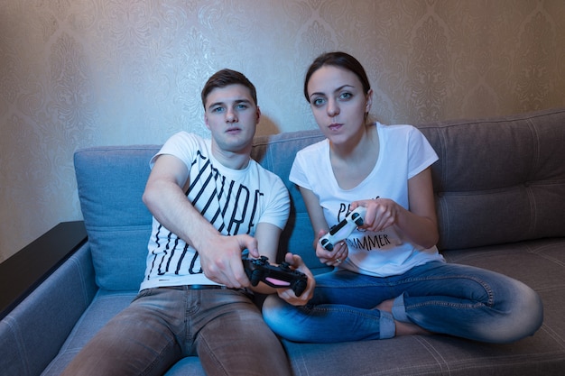 Vista frontal de una joven pareja sentada en un sofá juntos en casa jugando videojuegos con intensas expresiones competitivas