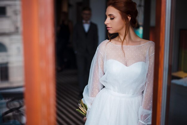 Foto vista frontal de una joven sensual en vestido de novia posando con ramo de flores fondo borroso de un hombre guapo en traje primera reunión de la novia y el novio